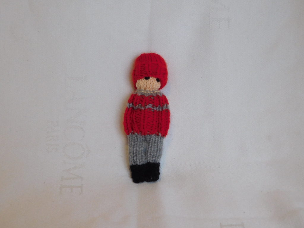 poupée garçon tricotée. Son patalon est gris, son pull est rouge et gris, son bonnet est rouge.